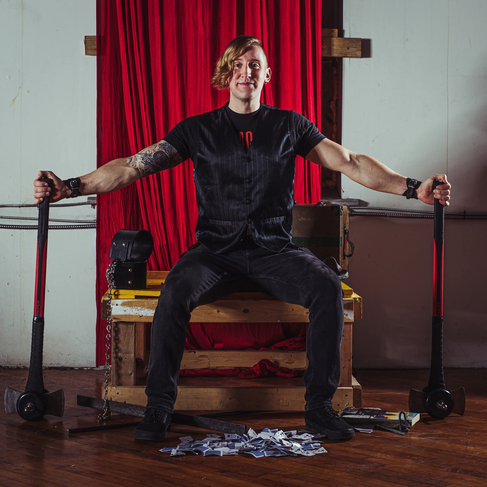 Professional portrait of Reid Wilson seated with bent steel props, red theatrical curtain background